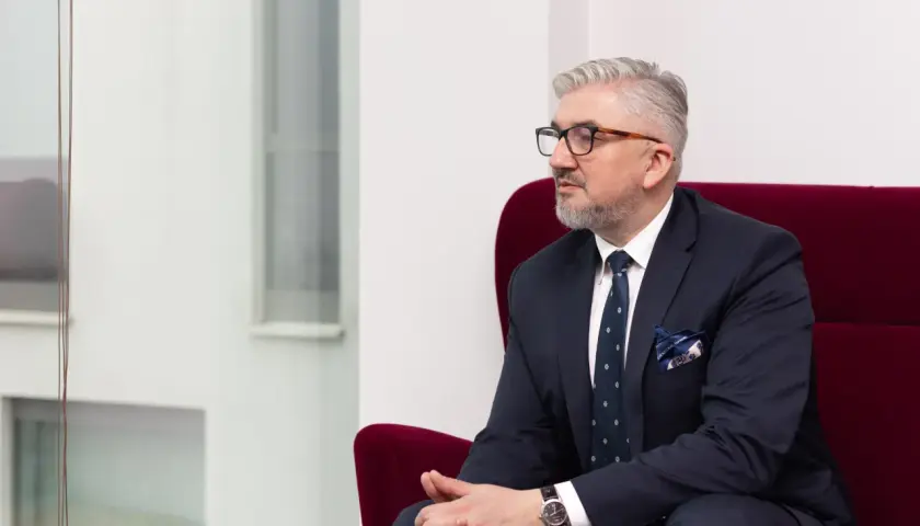 Mężczyzna w garniturze siedzi w czerwonym fotelu, patrząc w bok.