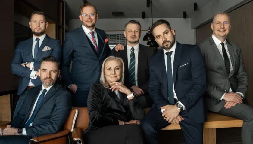 Grupa profesjonalistów w garniturach, siedząca i stojąca, w nowoczesnym wnętrzu.