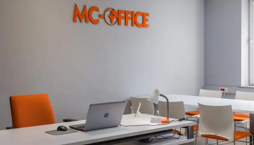 Pomarańczowe logo MC-OFFICE na szarej ścianie nad biurkiem z laptopem.